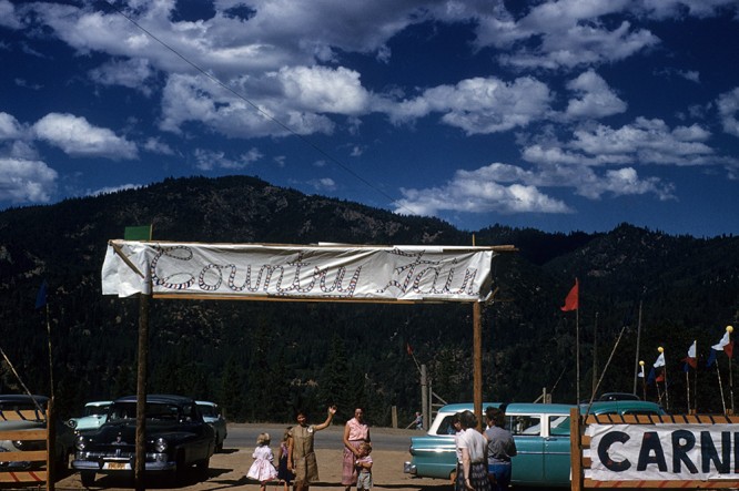 People stand under "Country Fair" sign in camp parking lot, 1950s 