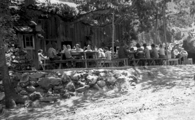 Black and white photo of campers enjoying a meal on the dirt floor eating platform, under large oak trees.